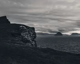 Far Peaks Black and white view of the cliffs at Trælanípa with distant islands under heavy clouds in the Faroe Islands.