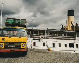 Quayside Cargo An old yellow Volvo truck loaded with crates stands beside a heritage steamboat on a cobbled quay under brooding skies.